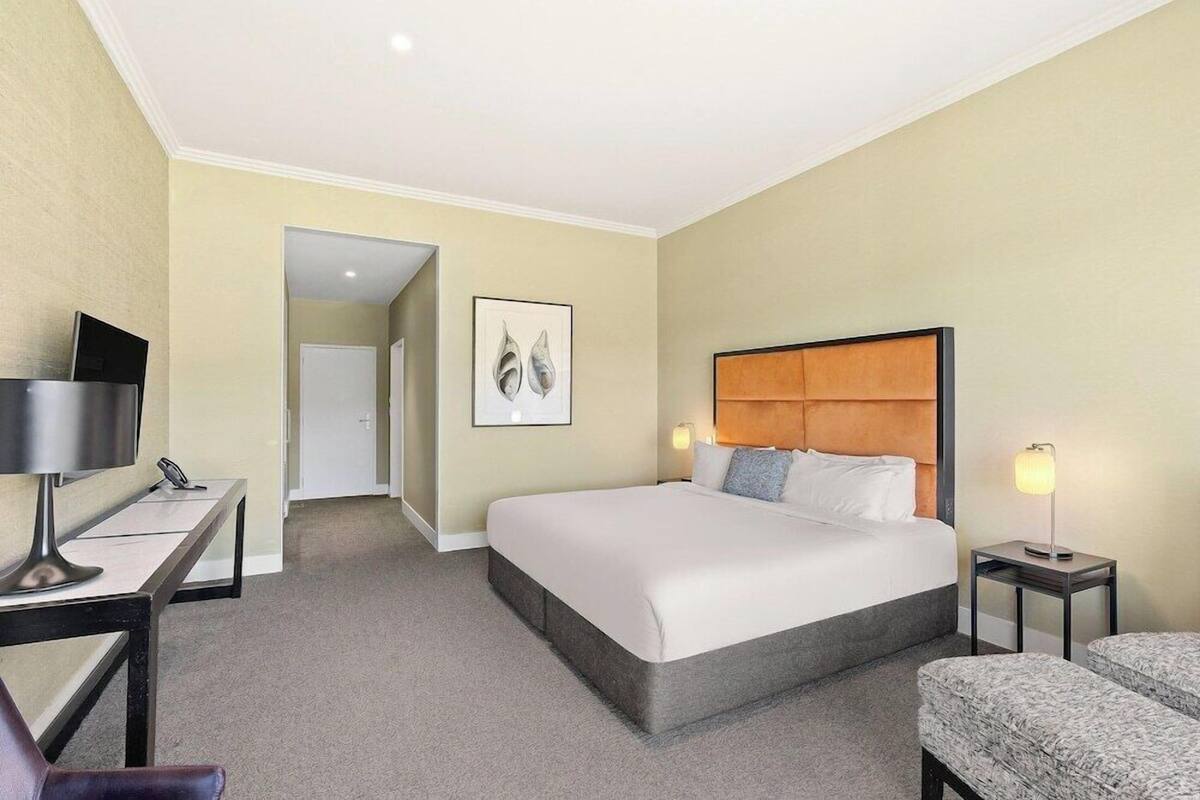 A hotel room with a bed, bedside tables, a desk, a chair, and a framed artwork on the wall.