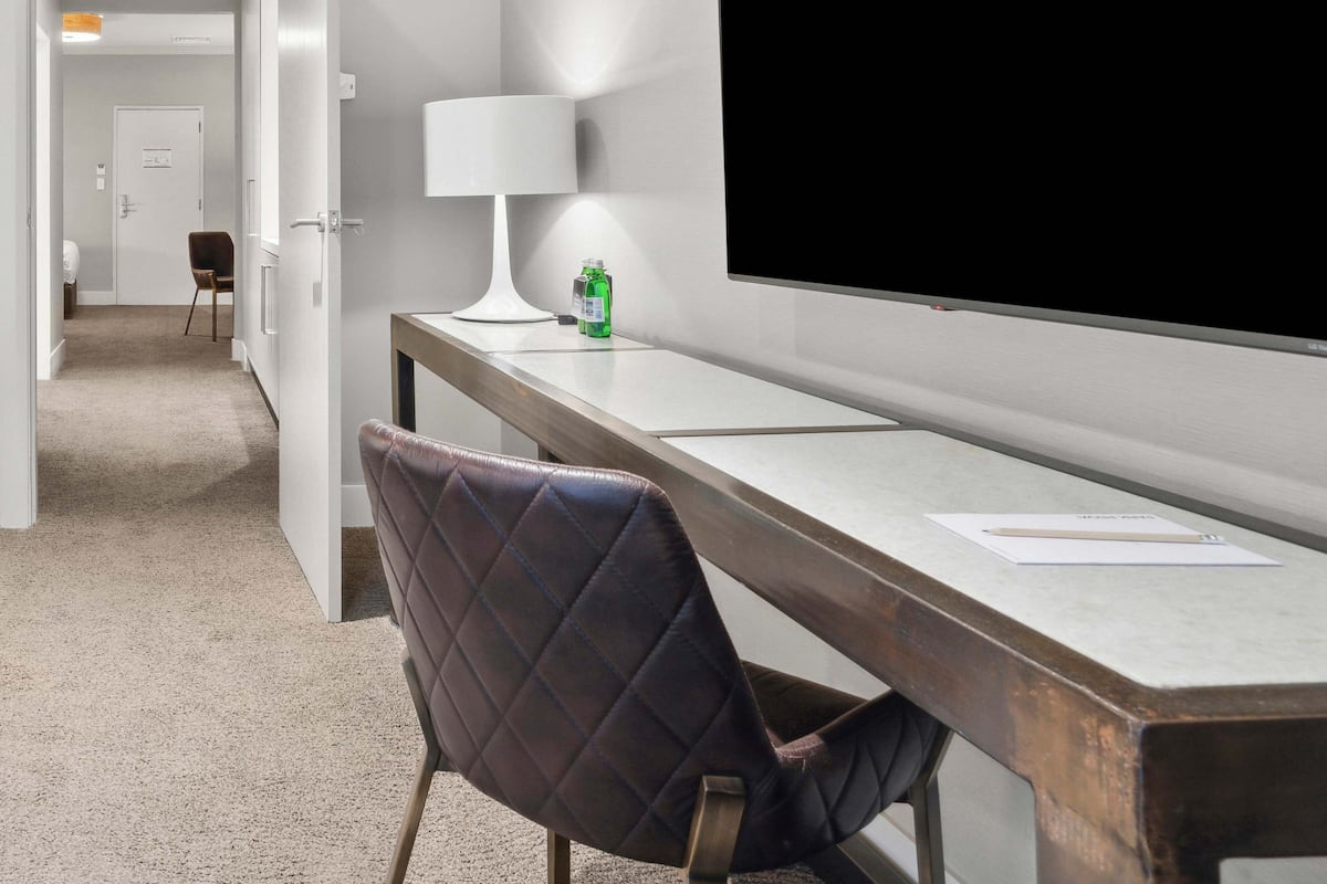A hotel room with a large flat-screen TV mounted on the wall, a desk with a chair, a lamp, and a bottle of hand sanitizer.