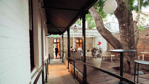 A covered outdoor seating area with tables and chairs, a tree, and a building entrance. - Little Drifter Surry Hills (Redfern)