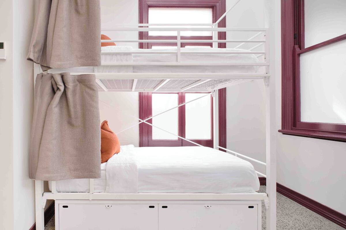 Een stapelbed met raam, een wit hoofdeinde en een bruin gordijn.