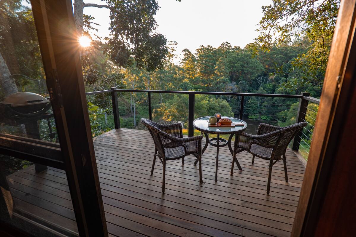 A wooden deck with wicker chairs and a table, overlooking a forested area.