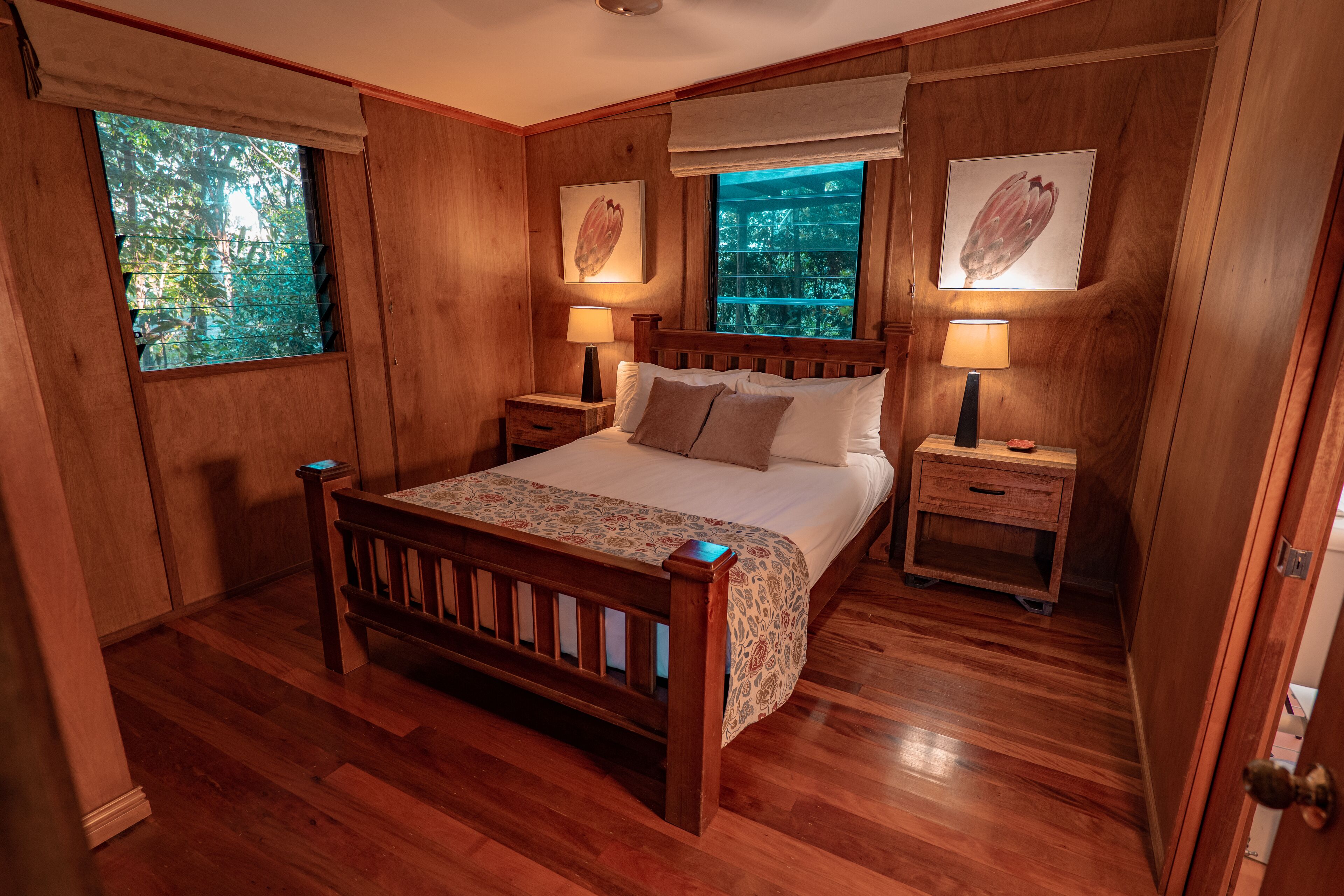 A wooden-paneled bedroom with a large bed, two bedside tables, and two windows.