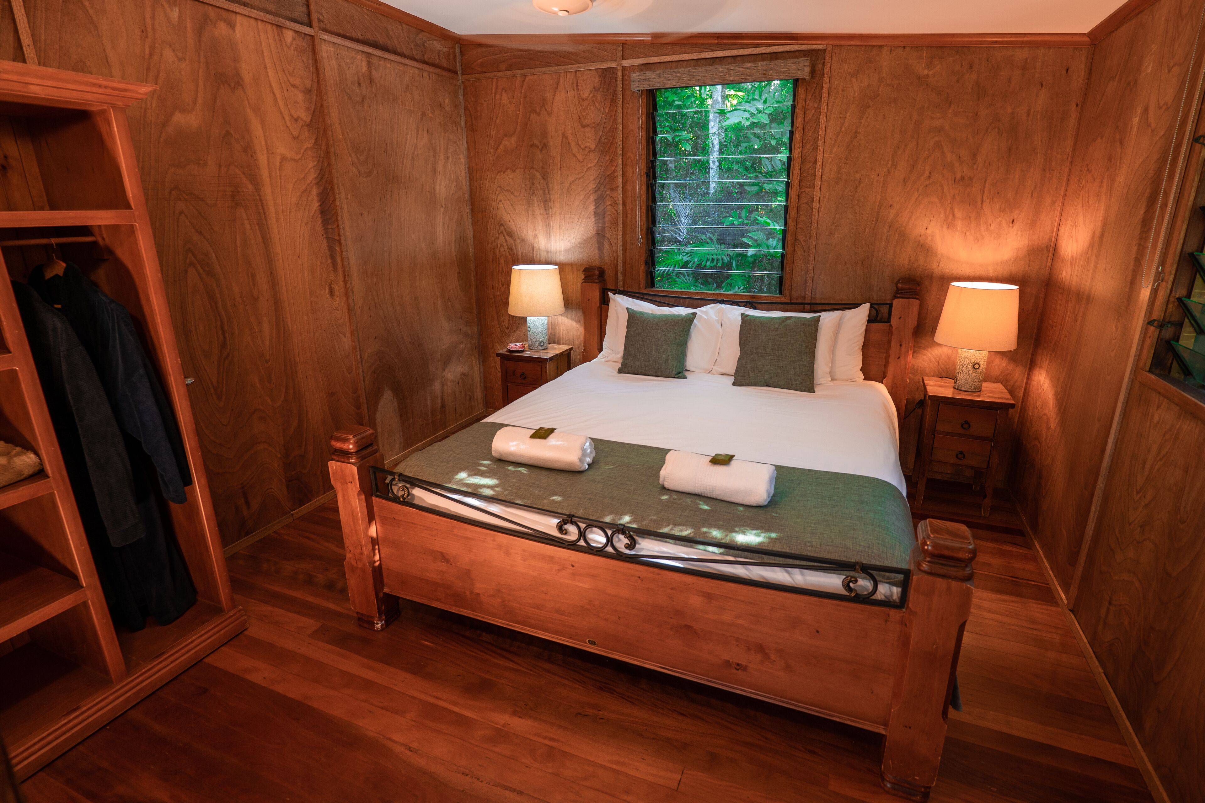 Une chambre à coucher aux murs lambrissés, avec un lit, deux tables de chevet dotées de lampes, une armoire en bois et une fenêtre donnant sur un paysage verdoyant.