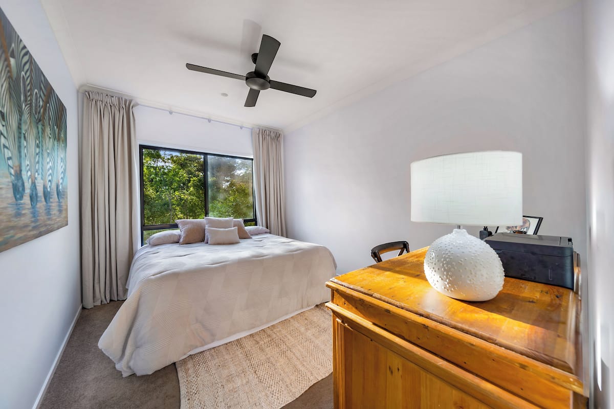 A bedroom with a bed, a ceiling fan, a window with curtains, a desk with a lamp, and a decorative item.