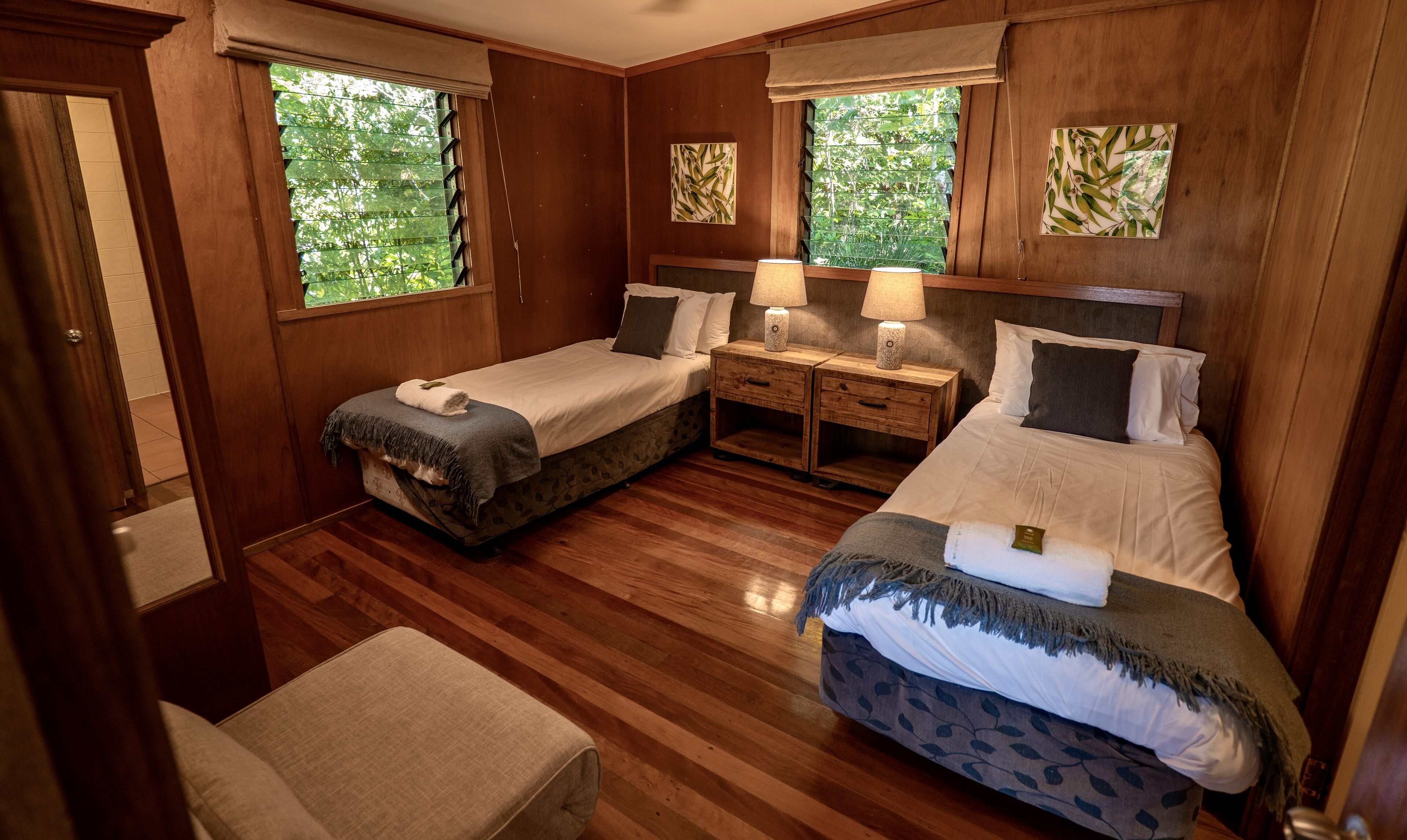 A room with two beds, a desk, and a chair. There are windows with views of greenery.