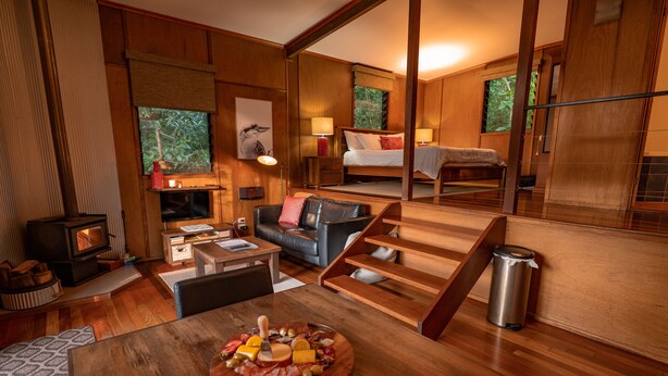 A modern, wood-furnished room with a fireplace, a dining area with a charcuterie board, a cozy seating area, and a bedroom with a large window. - Whispering Valley Cottage Retreat (Witta)