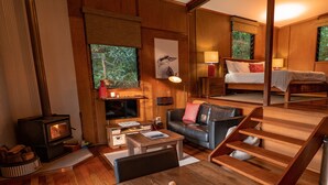 A cozy living room with a fireplace, a dining area with a fruit platter, and a bedroom with a bed and nightstands. - Whispering Valley Cottage Retreat (Witta)