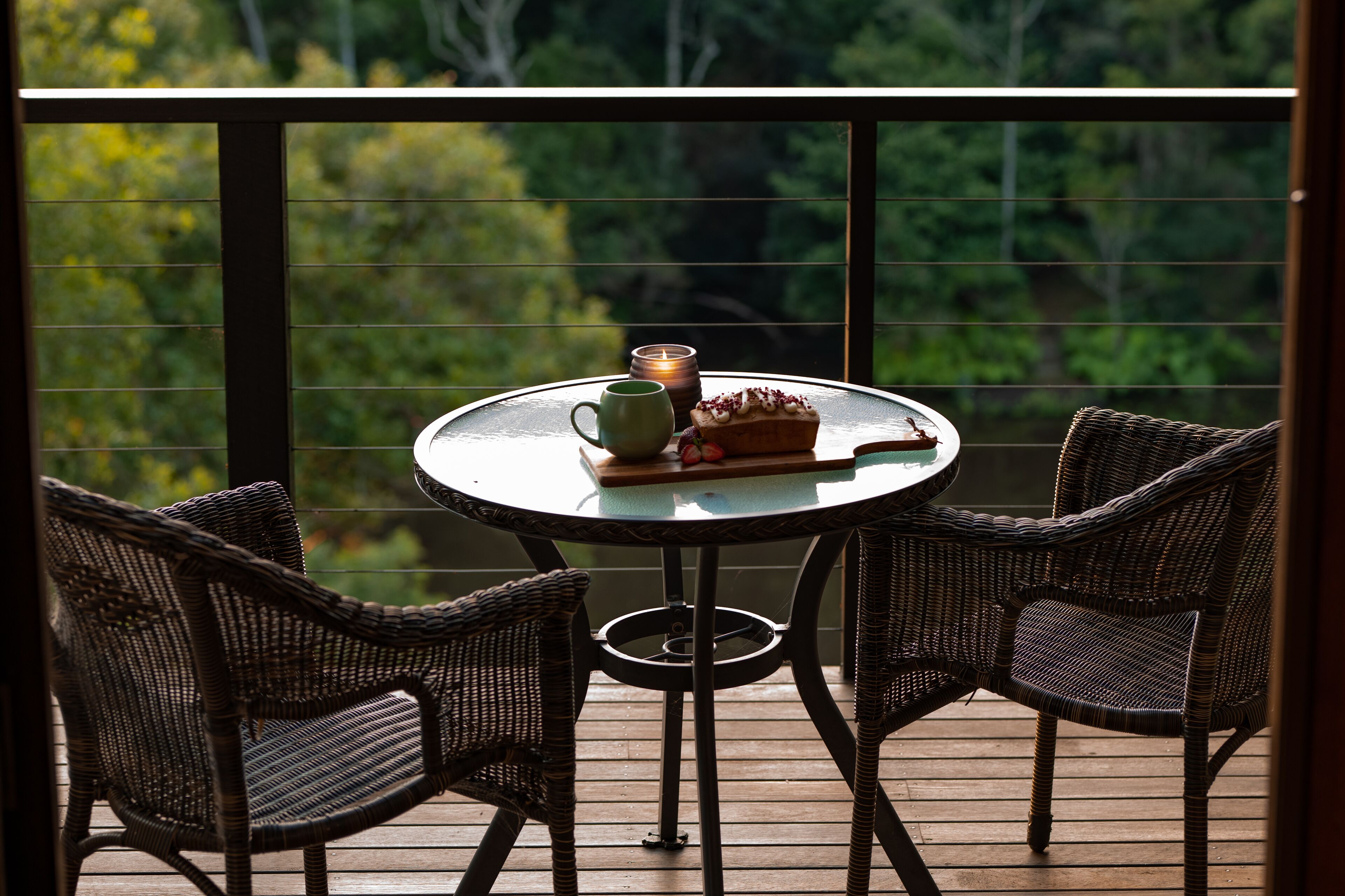A round table with two wicker chairs on a wooden deck, a candle, a mug, and a plate with food.