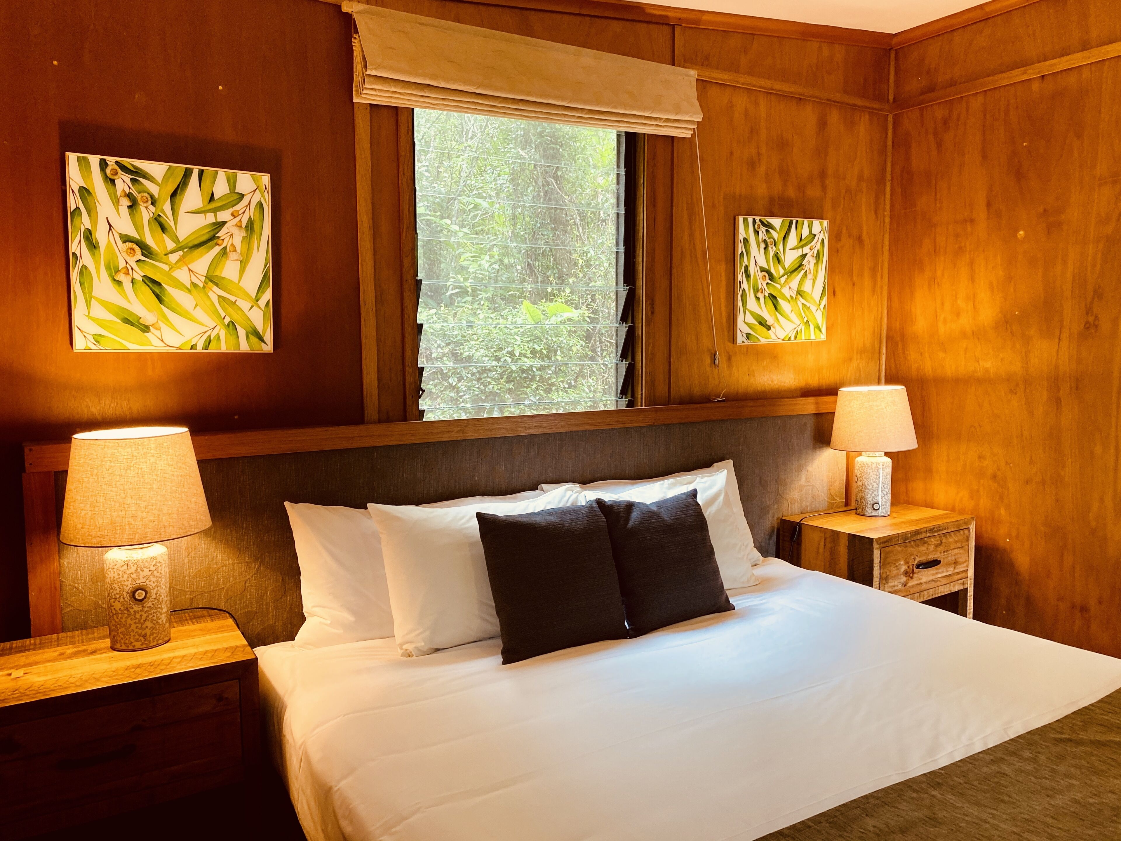 A neatly made bed with white and dark pillows, two bedside lamps, and a window with a view of greenery.