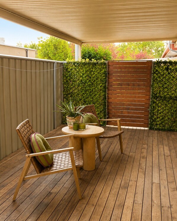An outdoor patio with a wooden table and chairs, a covered area, and a green hedge wall.
