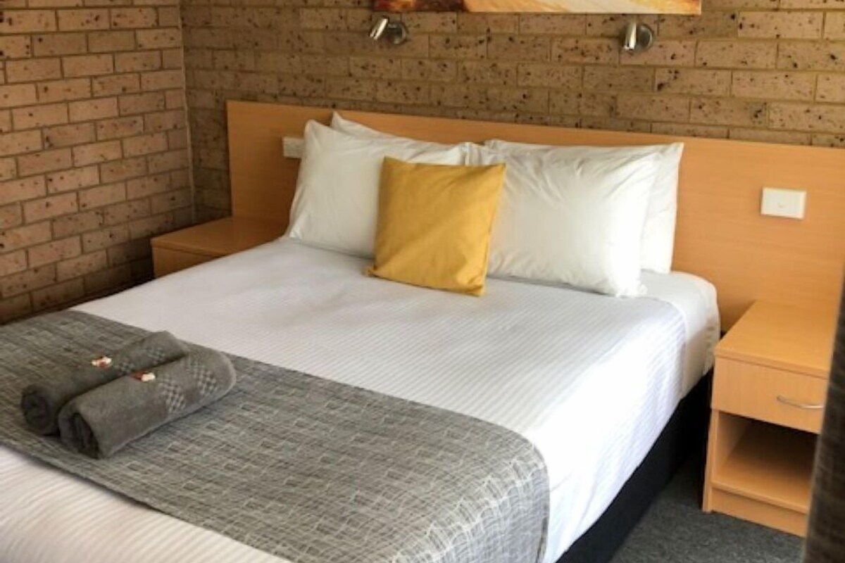 A hotel room with a brick wall, a bed with white and yellow pillows, a gray blanket, and a painting of a forest scene.