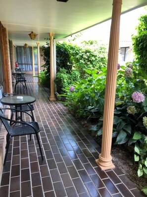 A covered outdoor patio with brick flooring, a table with chairs, and lush greenery.