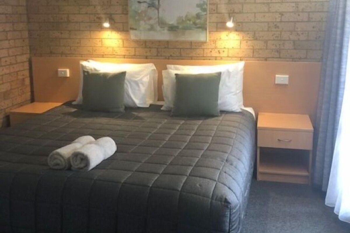 A hotel room with a brick wall, a large bed with a quilted cover, two bedside tables with lamps, and a painting of a tree.
