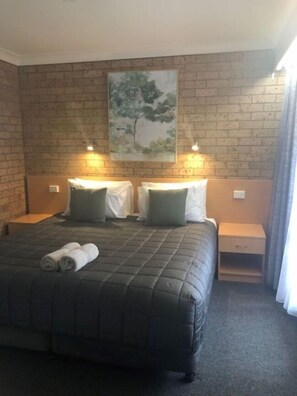 A hotel room with a brick wall, a large bed with a quilted cover, two bedside tables with lamps, and a painting of a tree.