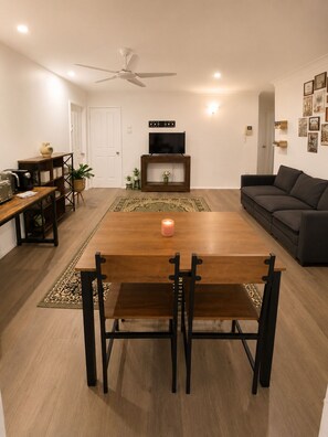 A modern living room with a wooden dining table, black chairs, a grey sofa, a TV on a wooden stand, and a ceiling fan. - Muswellbrook Motor Inn (Muswellbrook)