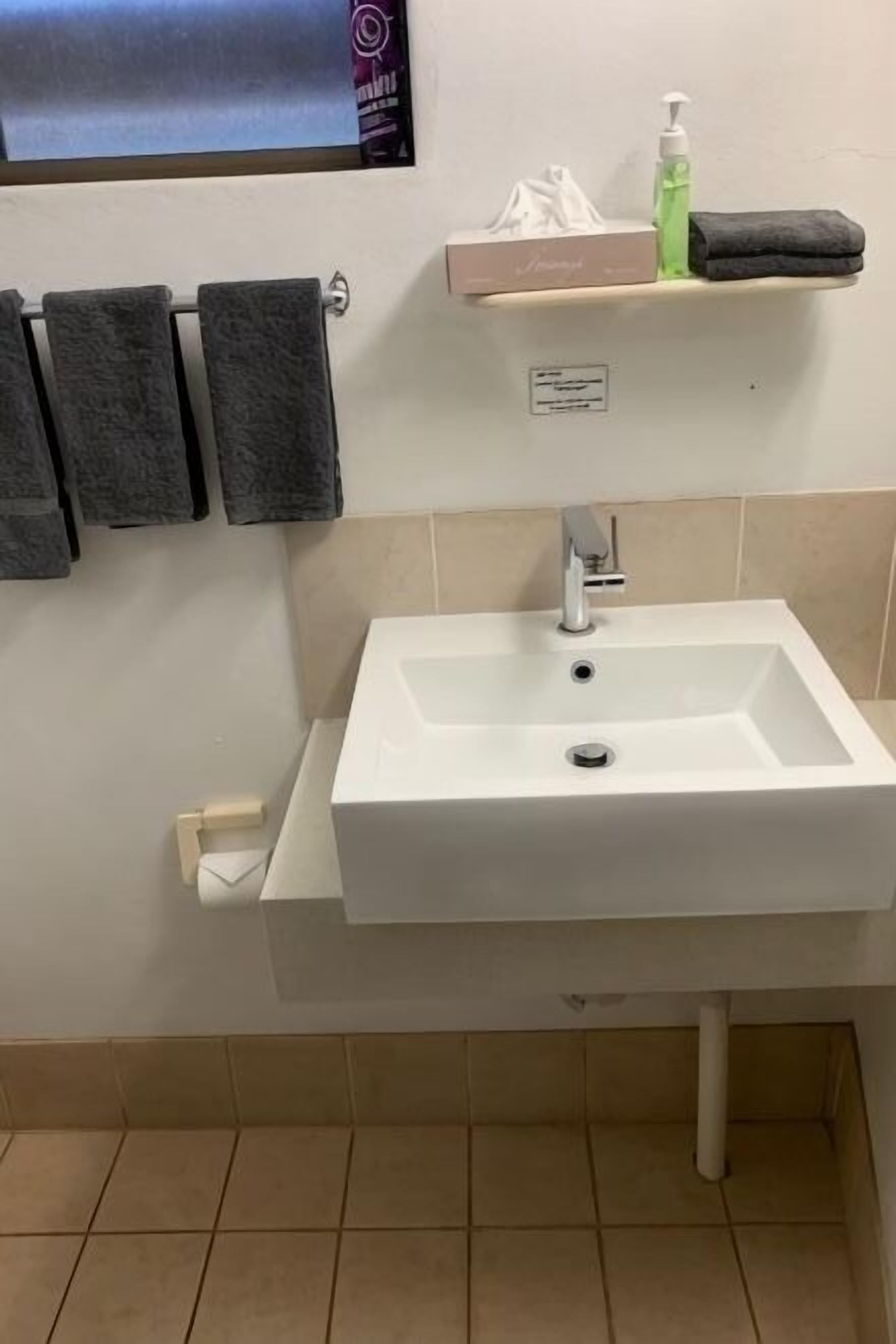 A bathroom with a white sink, a towel rack with three towels, a soap dispenser, and a small shelf with a tissue box.