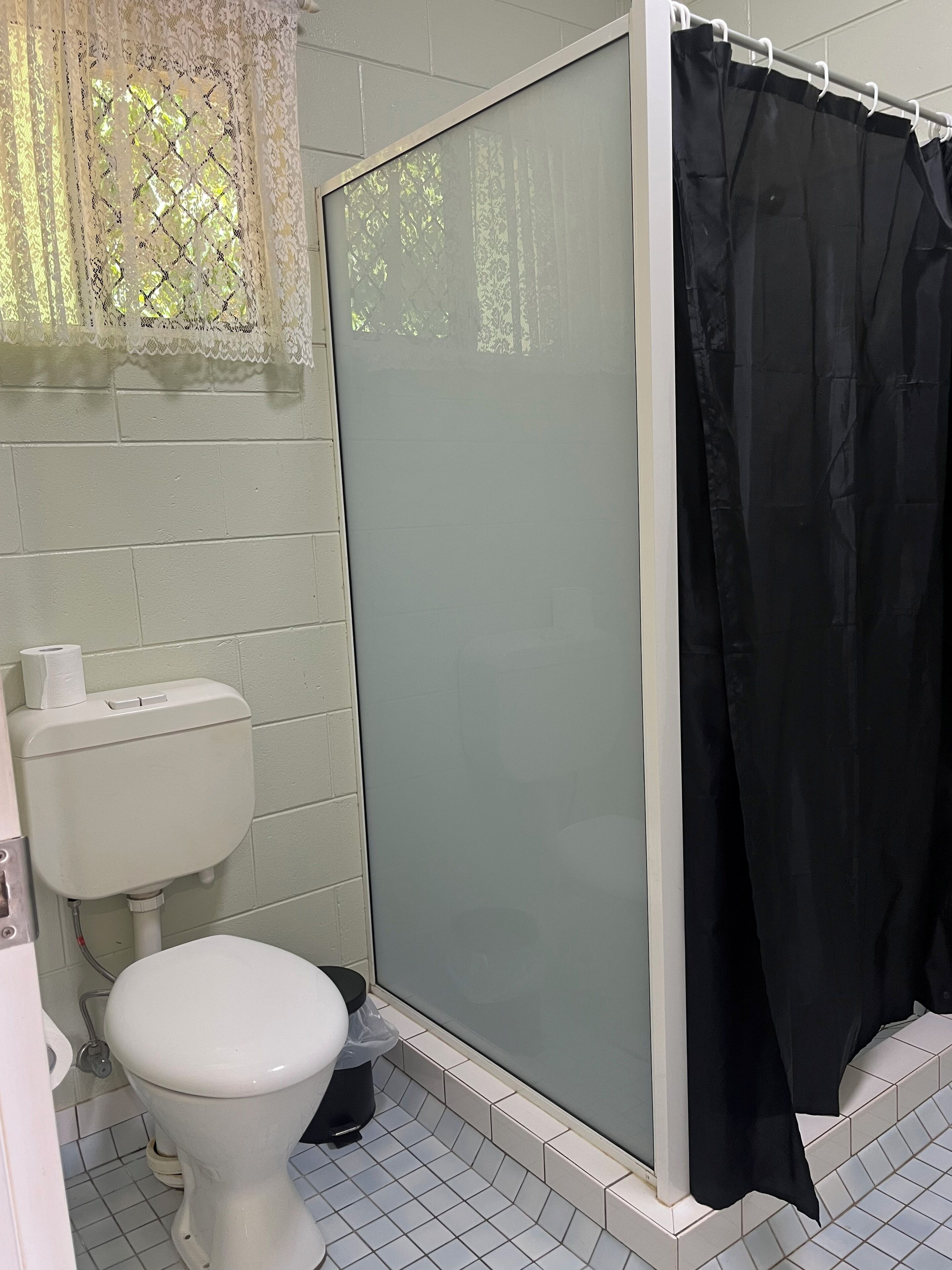 A bathroom with a toilet, a shower with a black curtain, and a lace window blind.