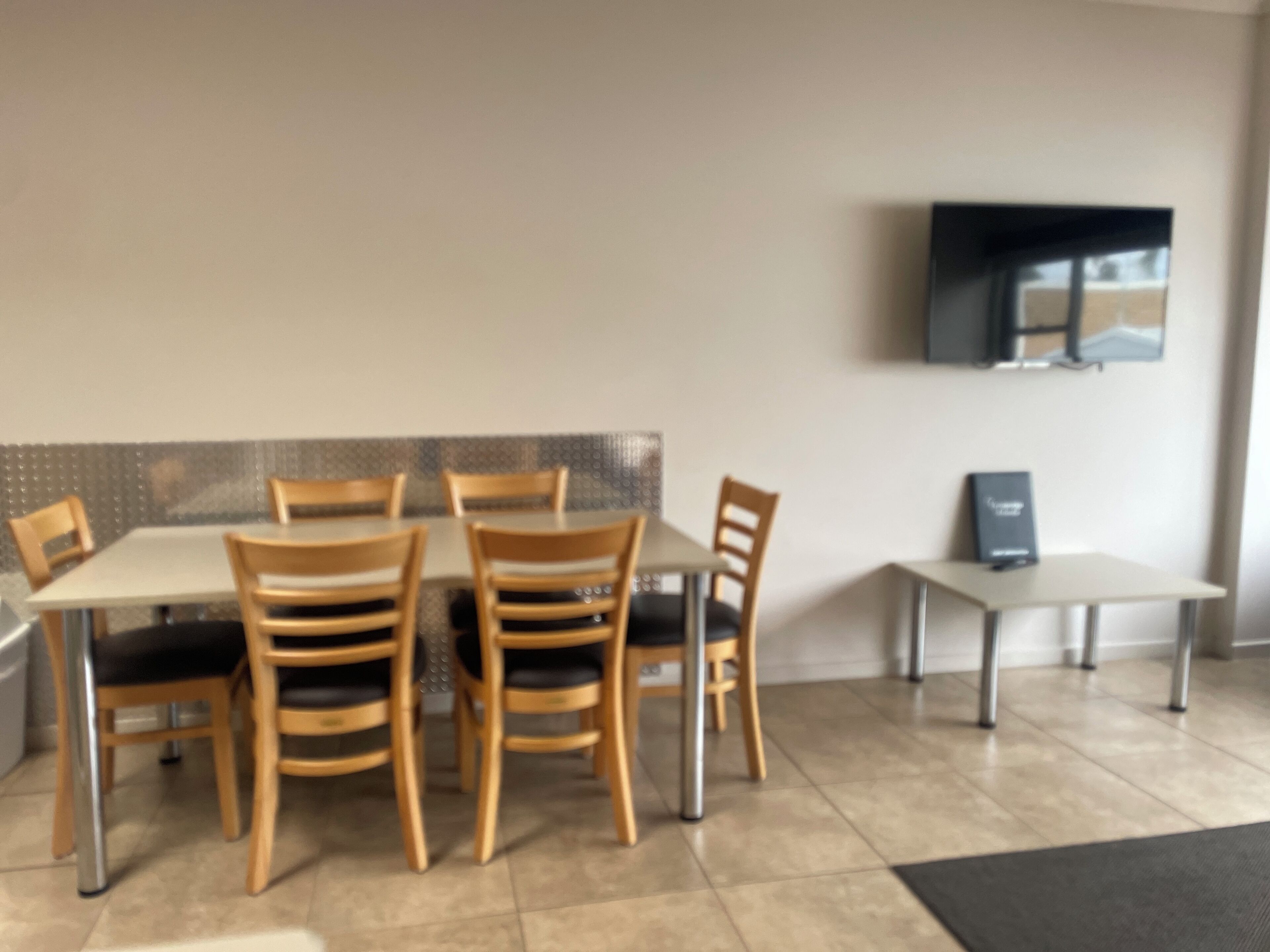A dining area with a table and chairs, a television mounted on the wall, and a small table with a book or menu.