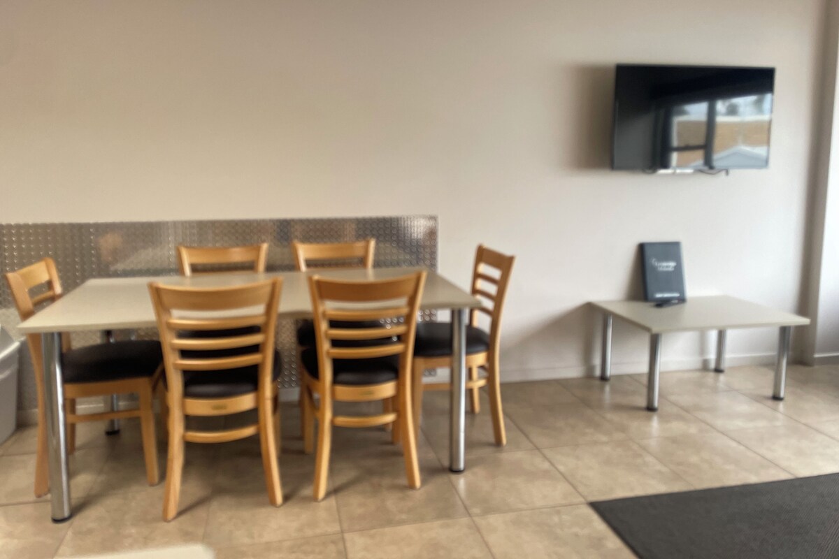 Uma área de refeições com mesa e cadeiras, uma televisão na parede e uma pequena mesa com um livro ou cardápio.