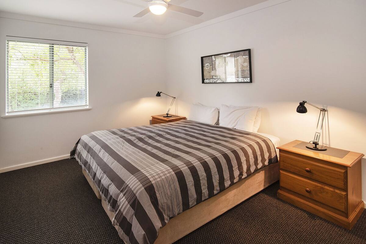 A bedroom with a bed, two bedside tables, a ceiling fan, a window with blinds, and a wall-mounted mirror.