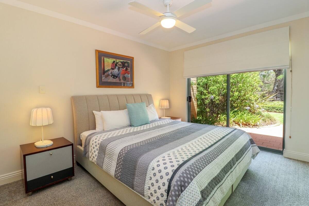 A bedroom with a bed, two bedside tables, a ceiling fan, a large window with a view of greenery, and a framed picture on the wall.