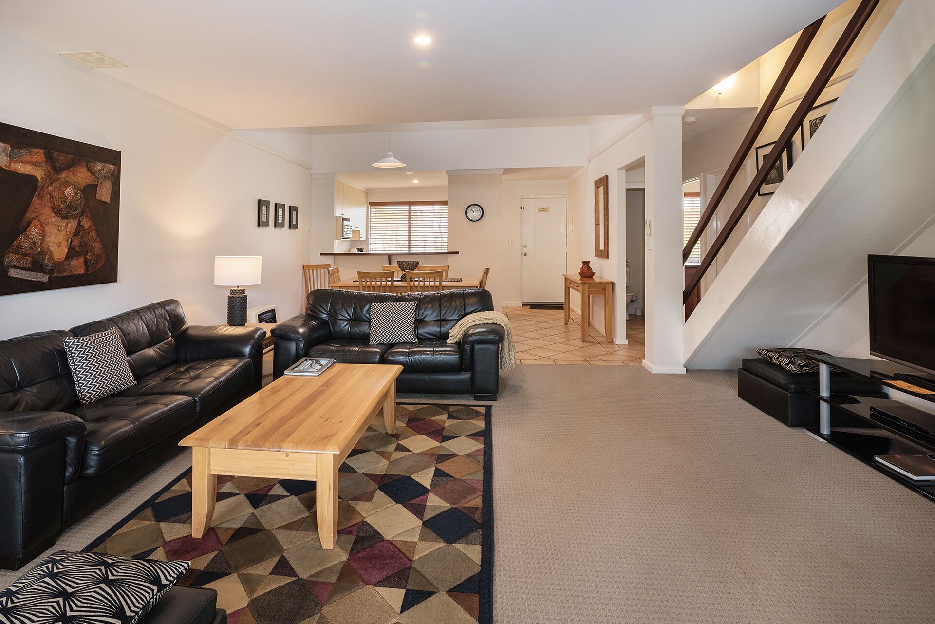 A modern living room with a black leather sofa, a wooden coffee table, and a patterned rug. There is a dining area in the background with a wooden table and chairs.