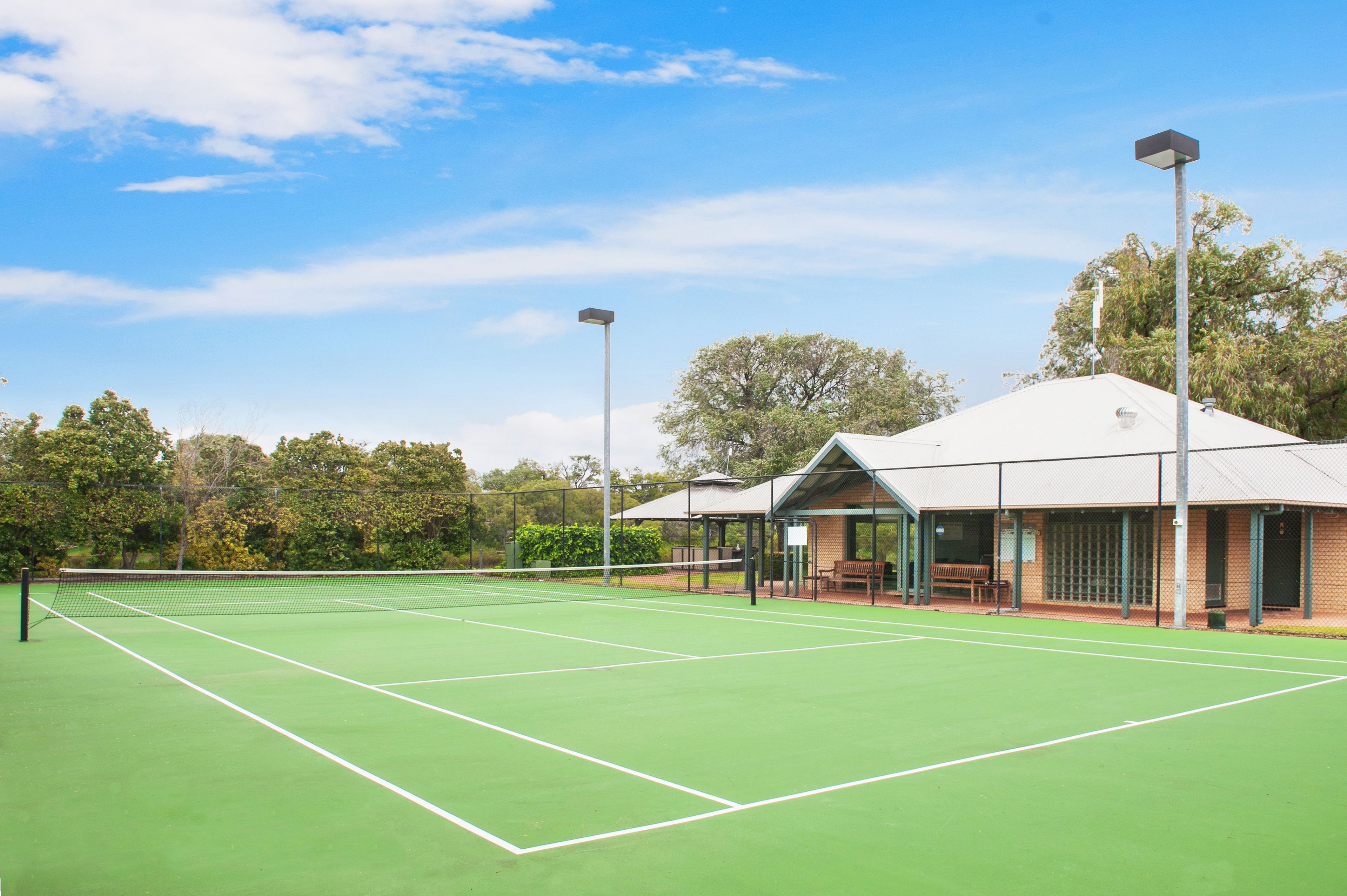 Una cancha de tenis al aire libre con un pabellón, bancos y árboles de fondo.