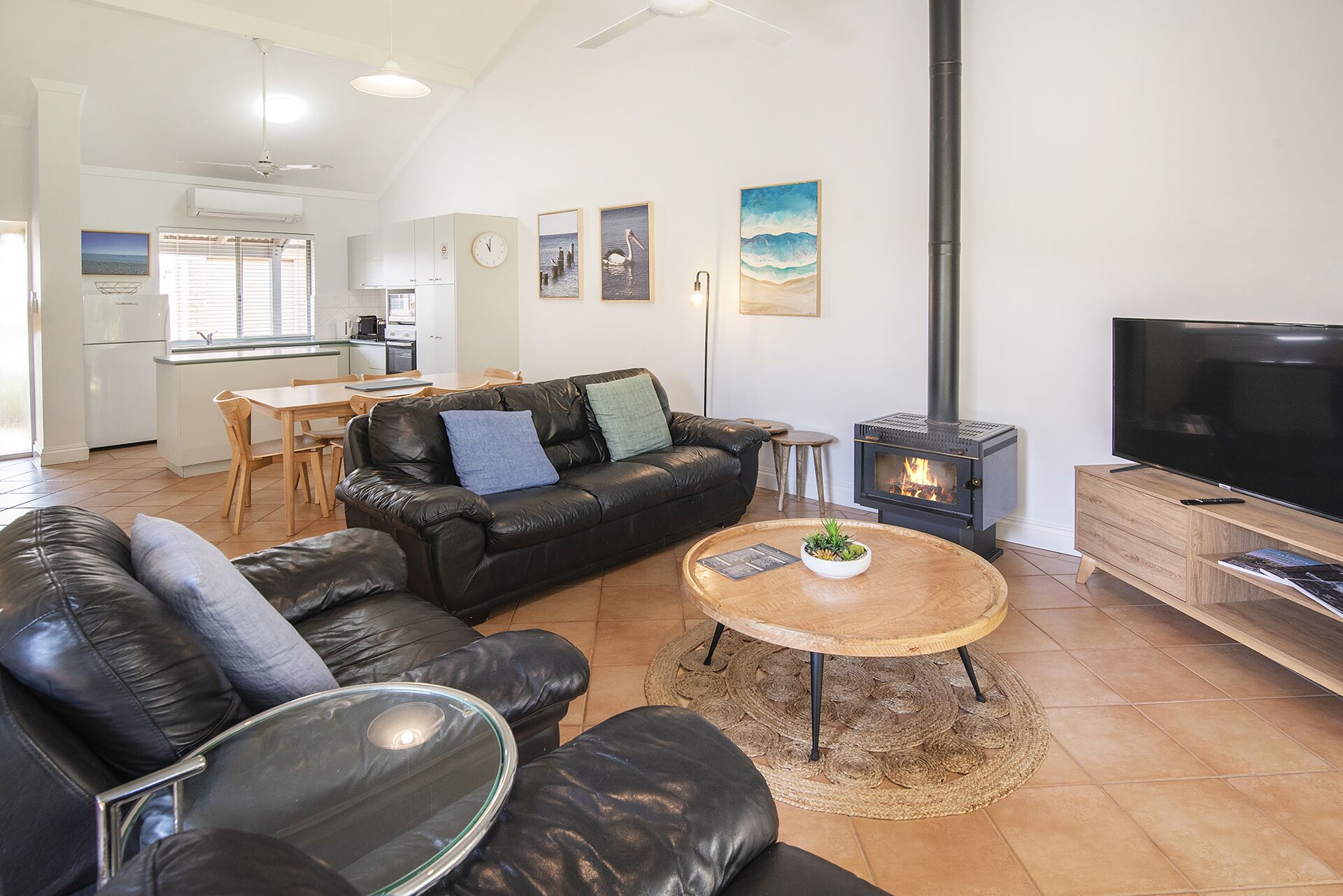 A modern living room with a black leather sofa, a wooden coffee table with a plant, a TV, and a wood-burning stove.