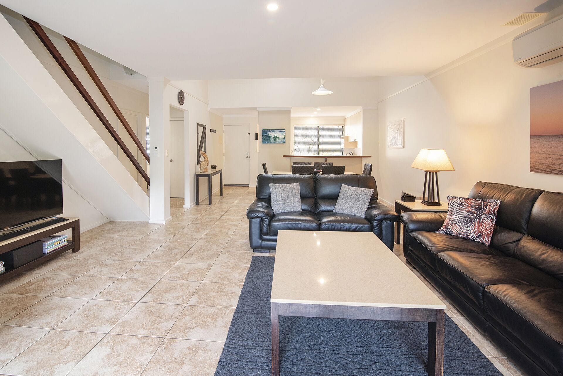 Una sala de estar moderna con un sofá, una mesa de centro y un televisor. Se ve una escalera a la izquierda y un comedor al fondo.