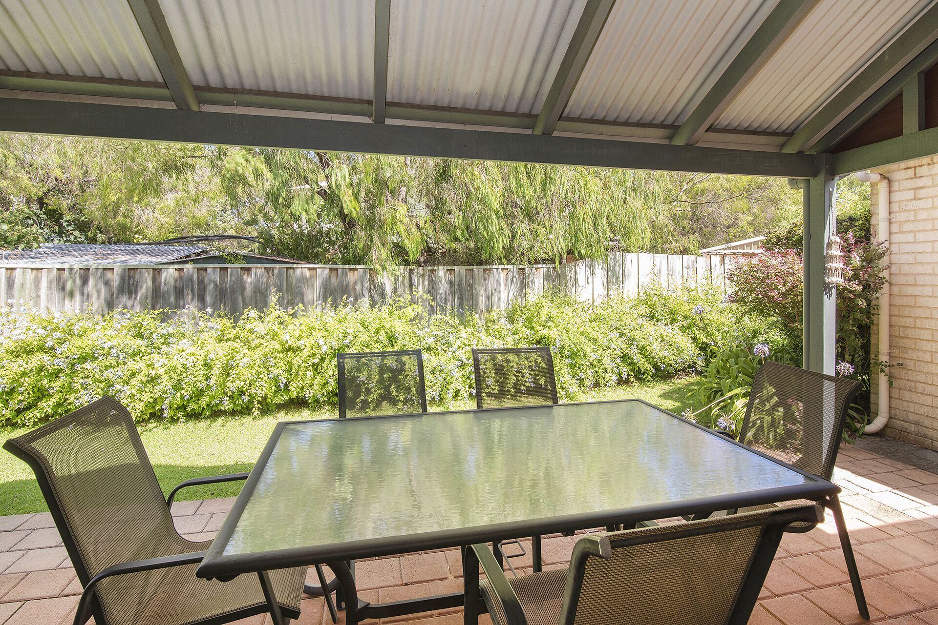 Un patio techado con una mesa de vidrio y cuatro sillas, rodeado de un jardín con diversas plantas y una valla de madera al fondo.