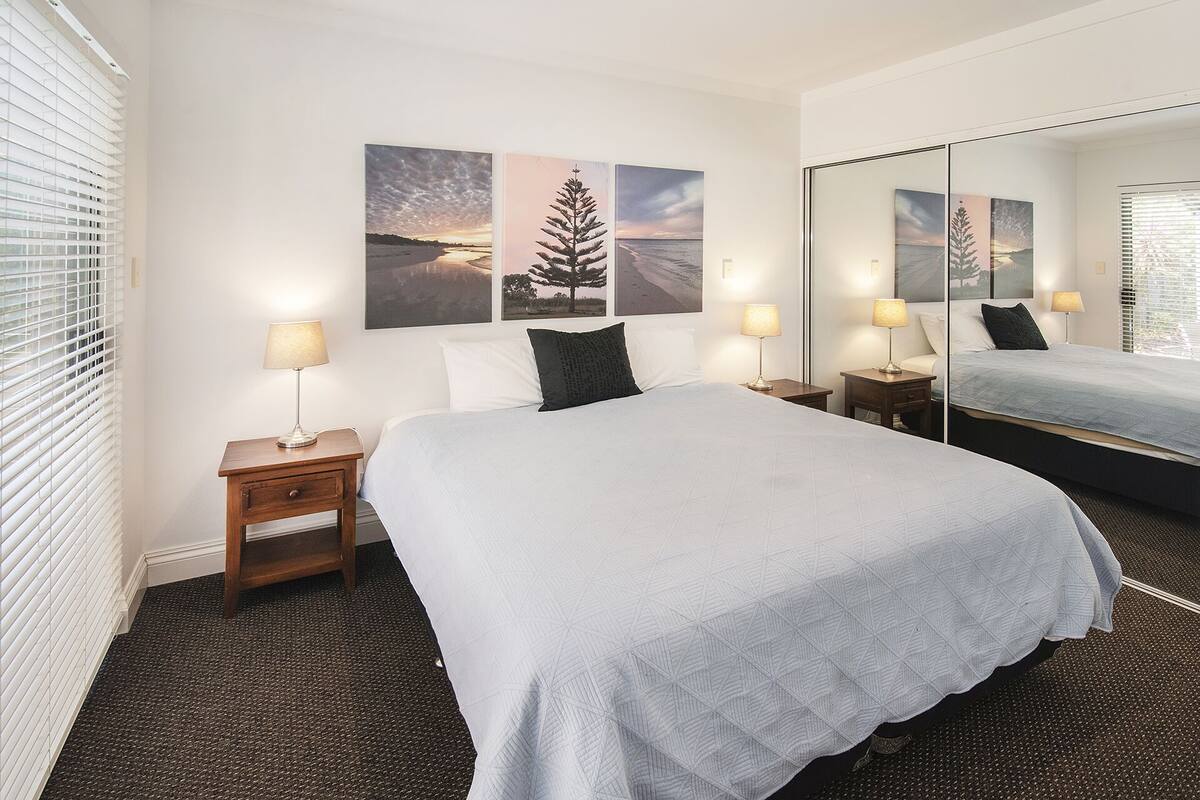 A hotel room with a large bed, two bedside tables with lamps, a mirror, and three landscape paintings on the wall.