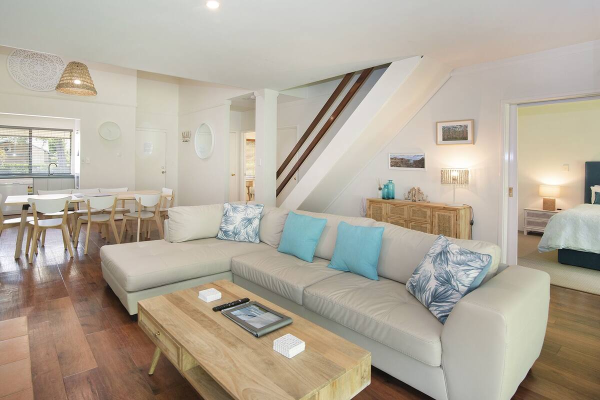 A modern living room with a large beige sectional sofa, a wooden coffee table, and a dining area with a white table and chairs.