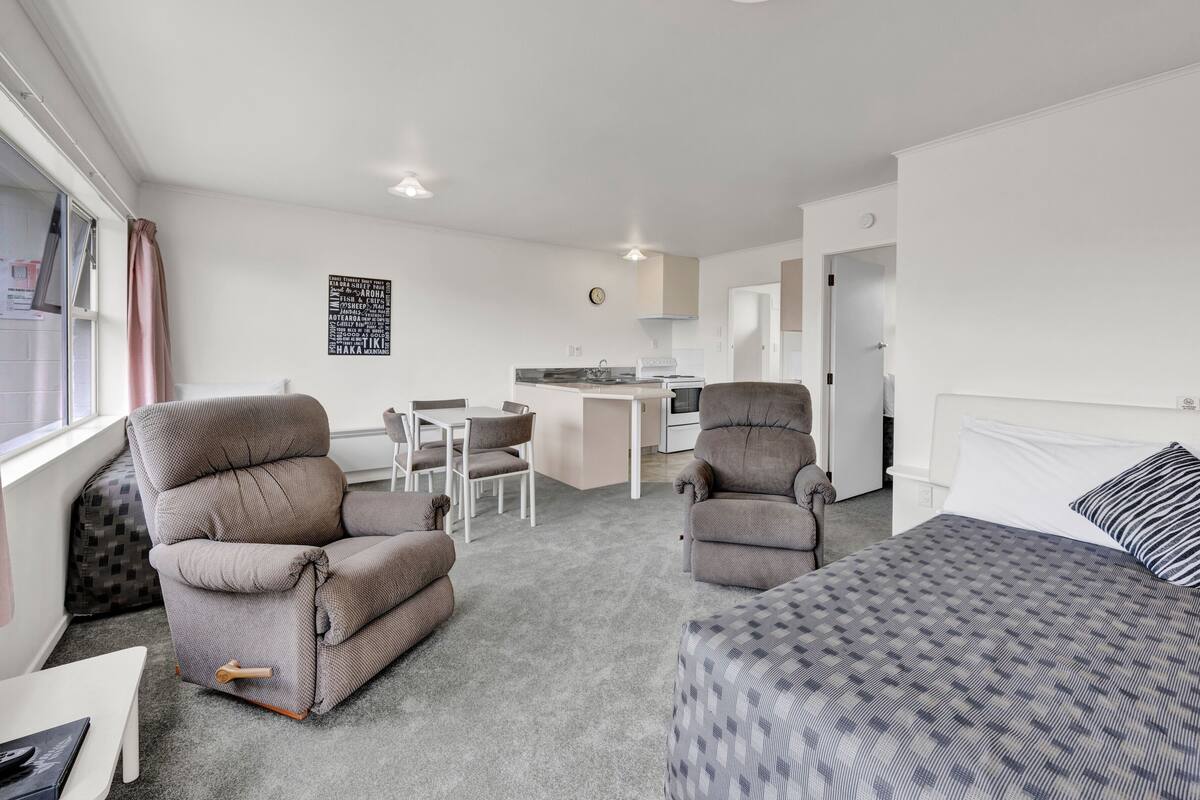 Une chambre d’hôtel compacte comprenant un coin cuisine, deux fauteuils, une table à manger et un lit avec une literie à motifs.