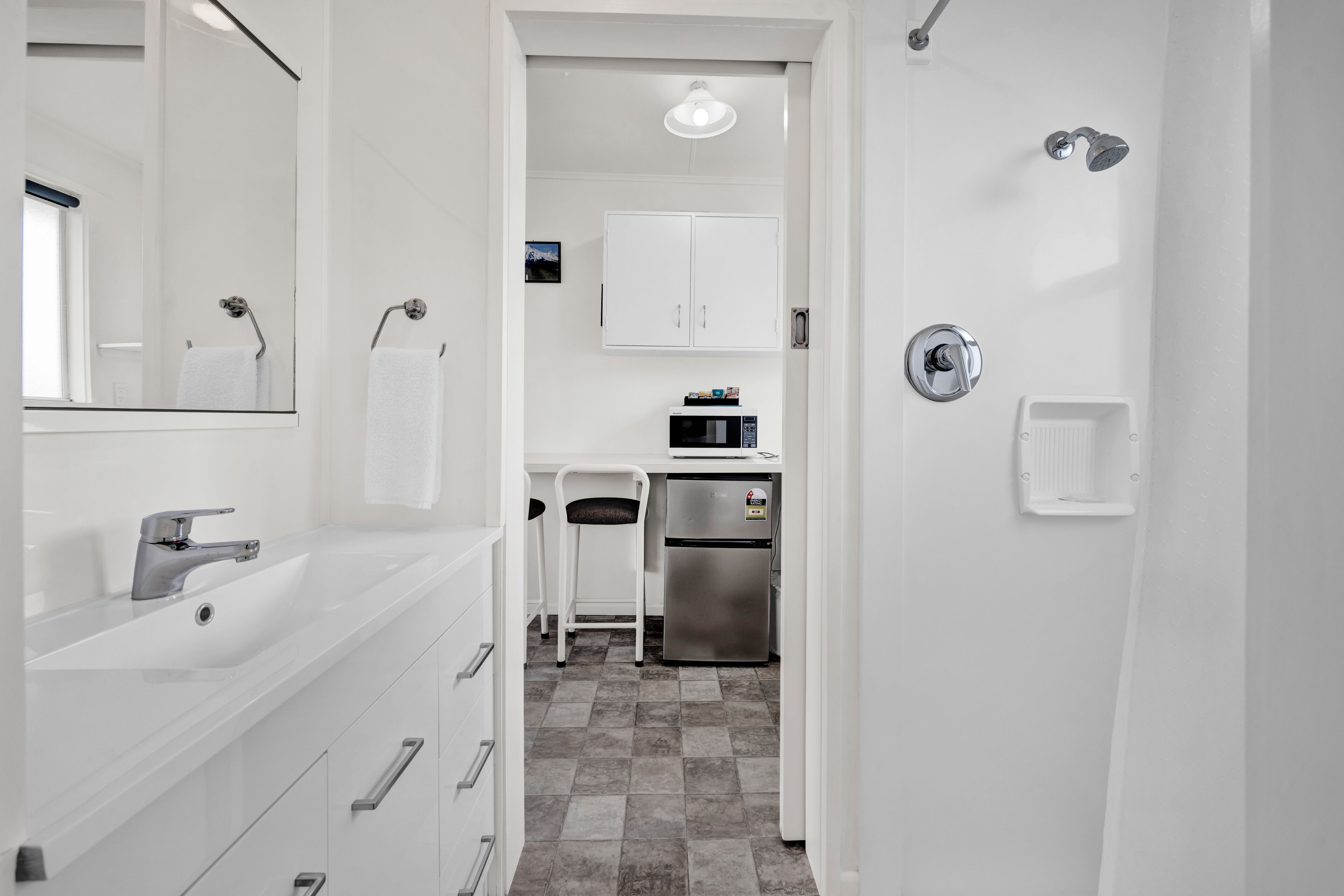 Un baño moderno con lavamanos, espejo y retrete blancos. Cuenta con una zona de ducha con cabezal de ducha de mano y un dispensador de papel higiénico en la pared.