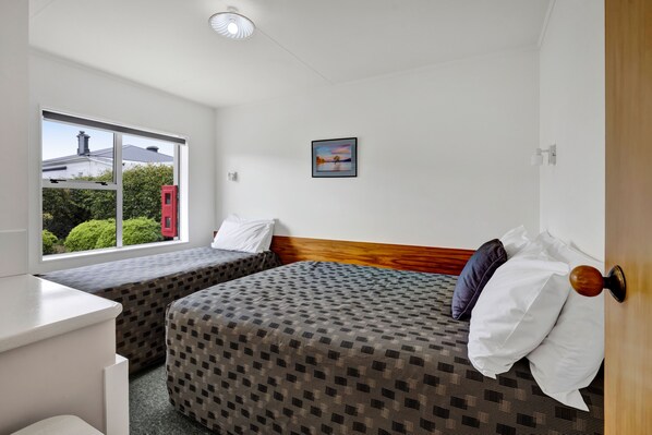 A hotel room with two single beds, a small desk, a window with a view of greenery and a building, and a framed picture on the wall. - Avon Motel (Hawera)