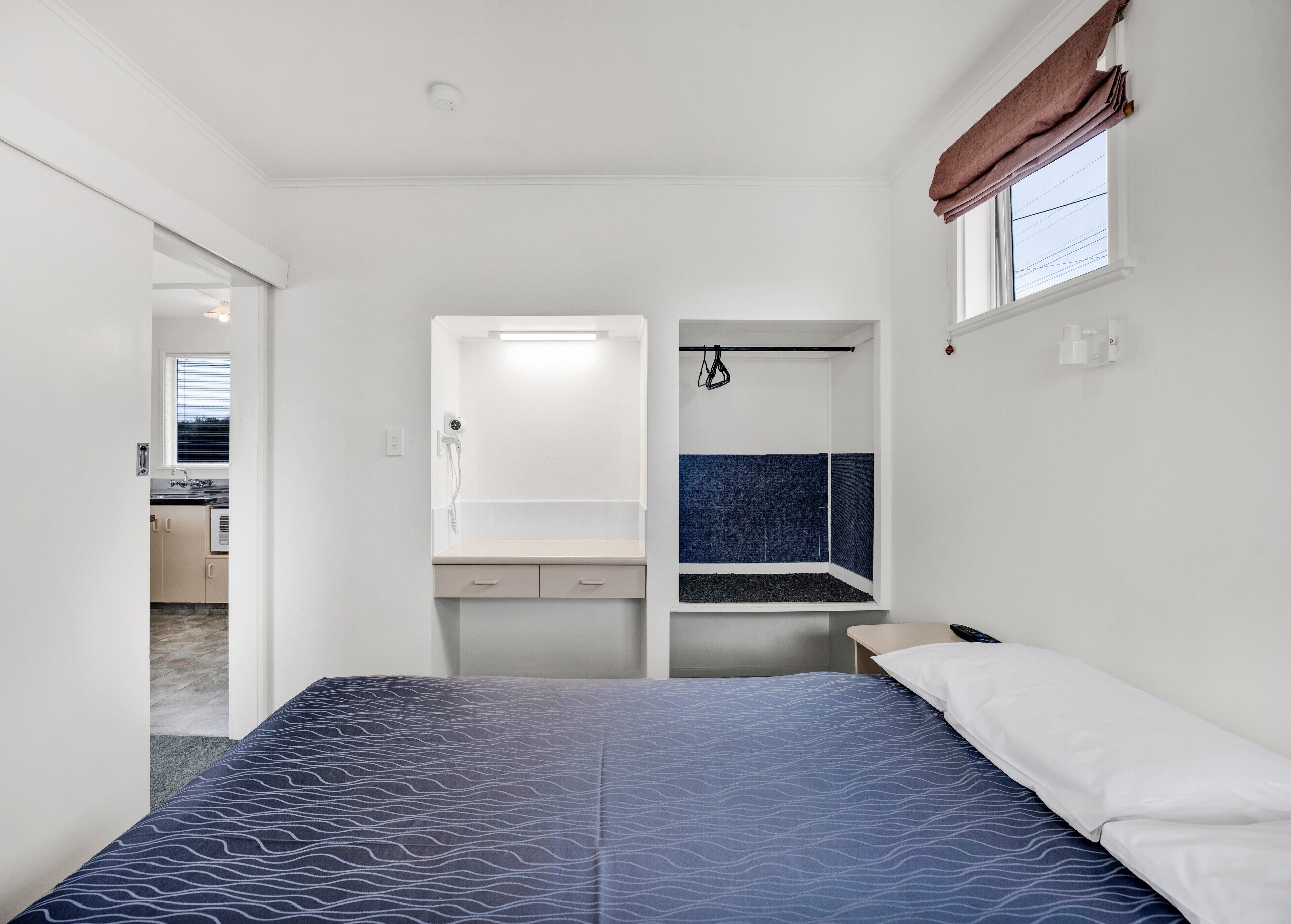 Un dormitorio con cama, un banco empotrado, un ventanal con cortinas y vista a una cocina.