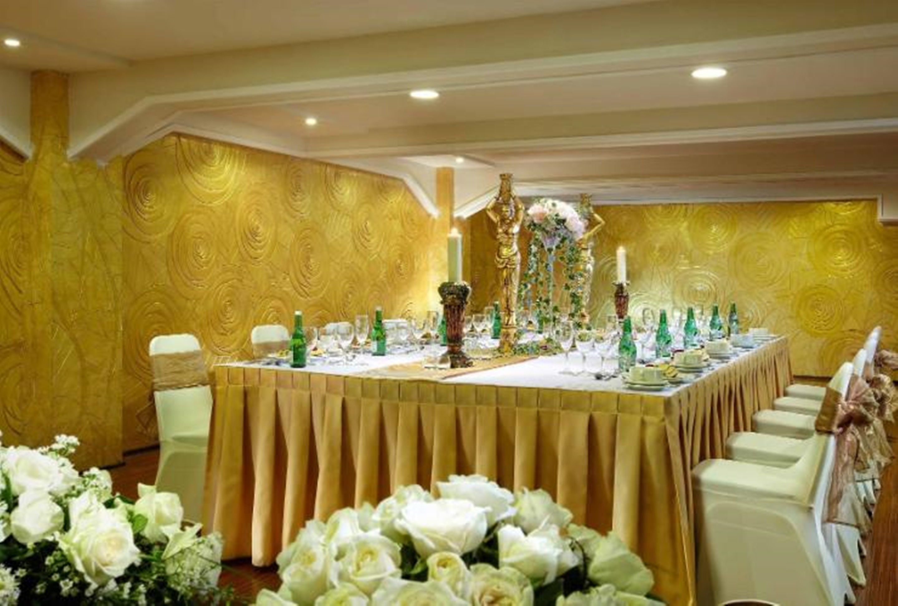 A banquet hall with a long table set for a formal event, surrounded by white chairs with gold sashes.