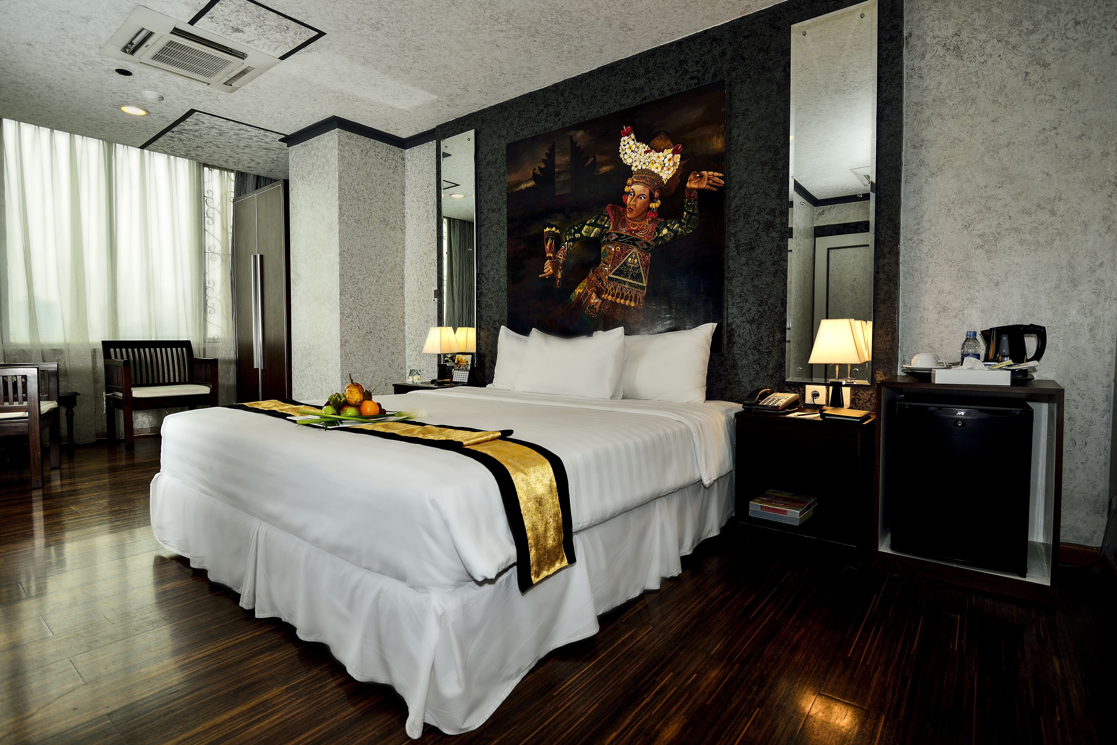 A large bed with white linens and a gold accent, a nightstand with a lamp, a small table with a coffee maker, and a large painting on the wall.