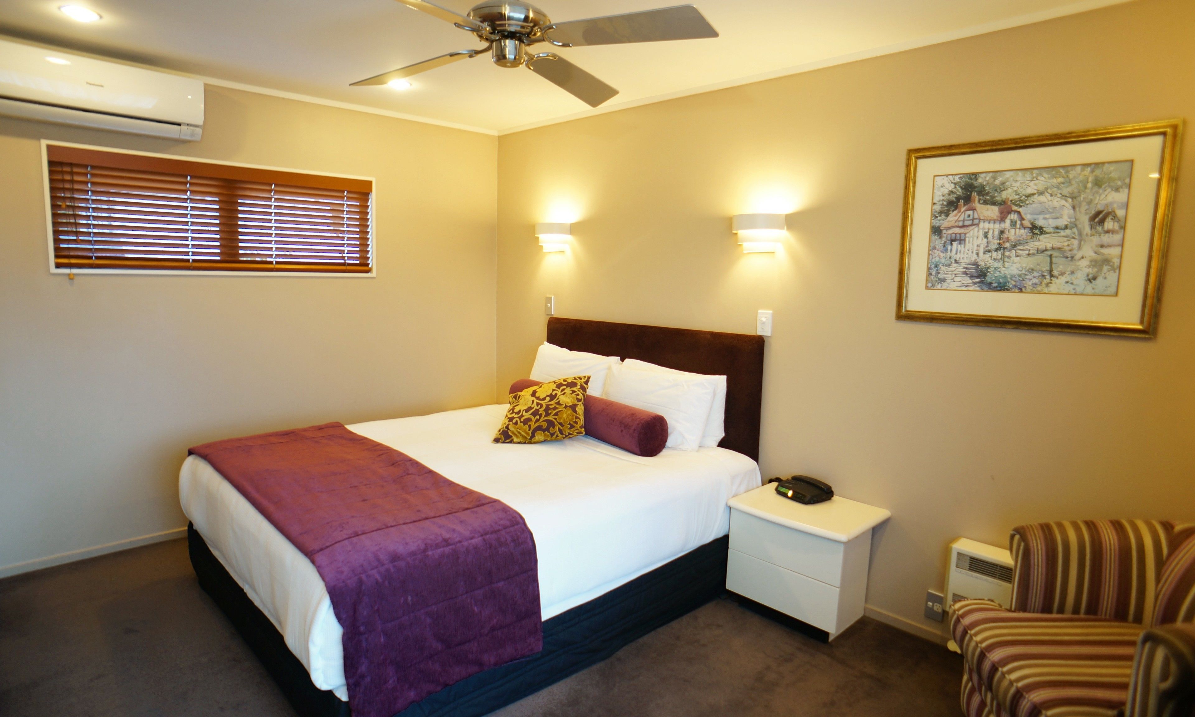 Una habitación de hotel con cama, mesita de noche, silla y ventilador de techo.