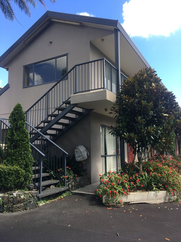 A two-story building with a staircase leading to a balcony, surrounded by greenery and flowering plants.