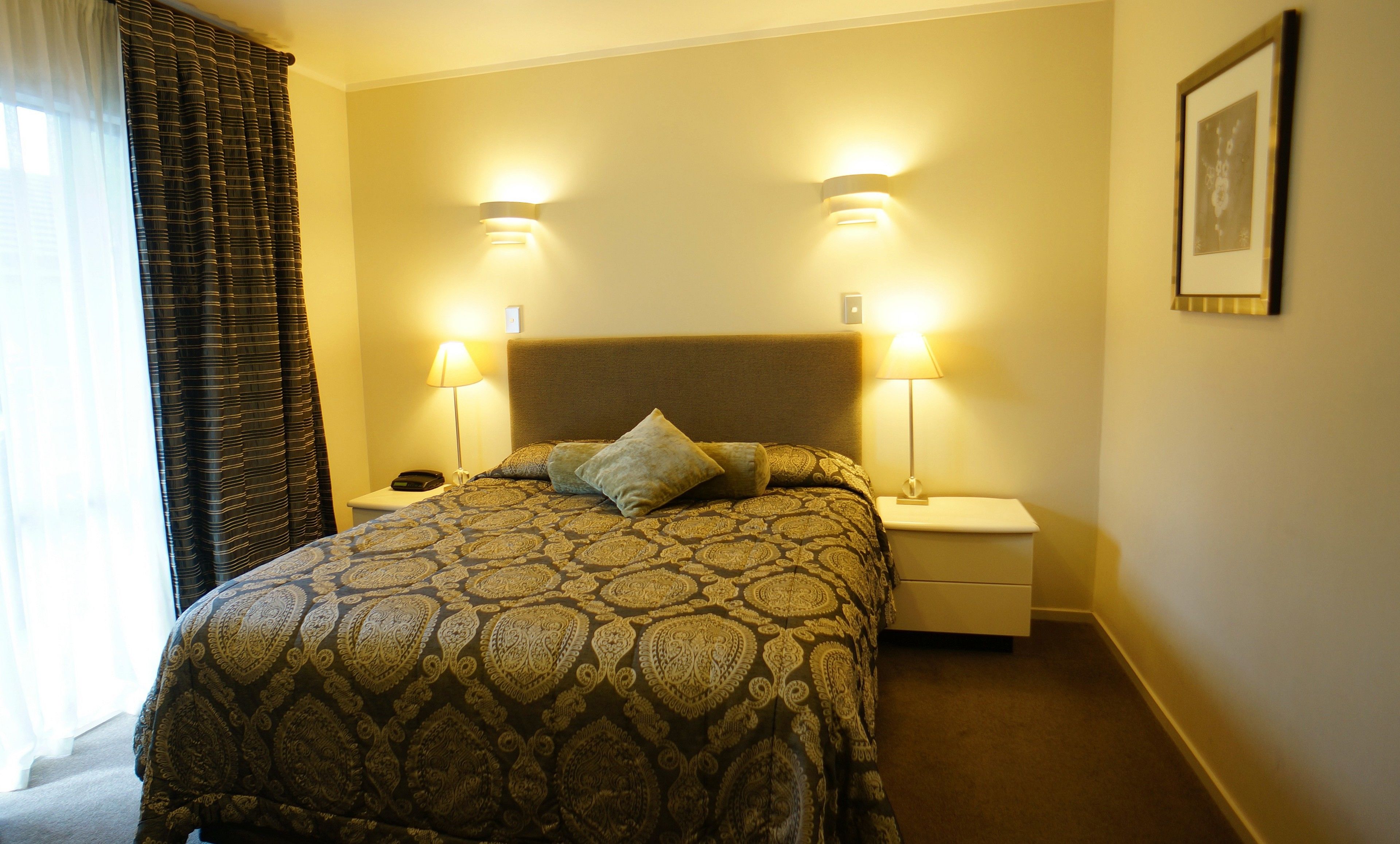 Ein Hotelzimmer mit einem gemusterten Bett, zwei Nachttischlampen, einem Nachttisch und einem gerahmten Bild an der Wand.