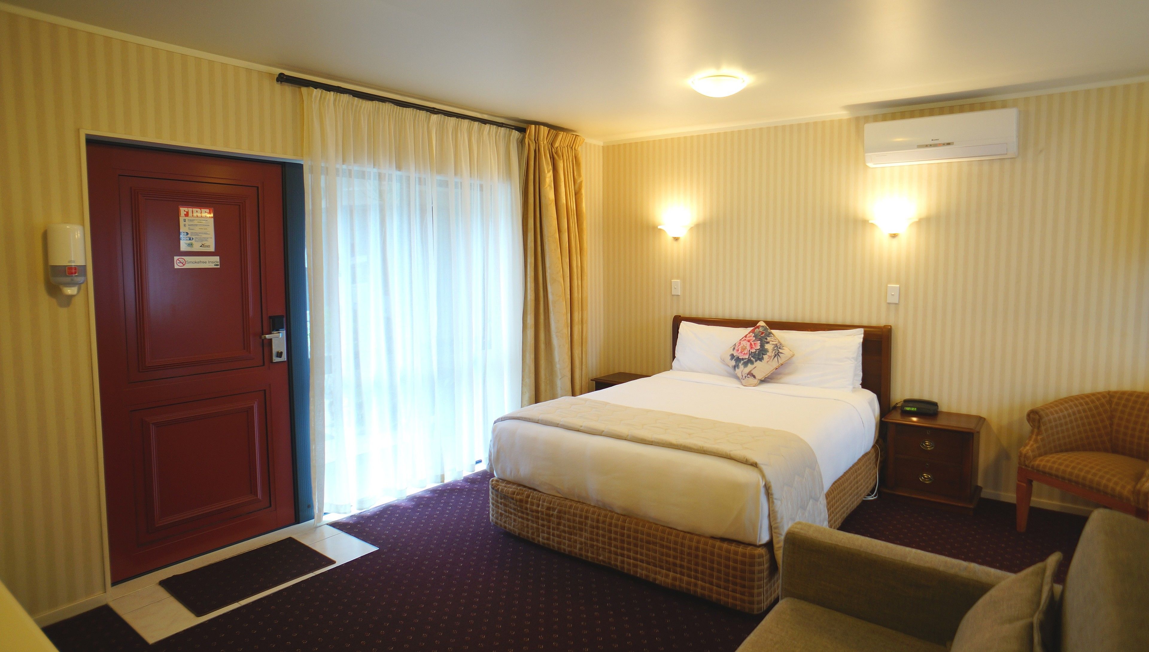 A hotel room with a bed, a chair, a nightstand, an air conditioning unit, and a door with a fire safety sign.