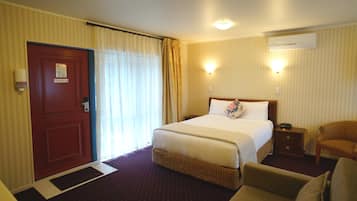 A hotel room with a bed, a chair, a nightstand, an air conditioning unit, and a door with a fire safety sign.