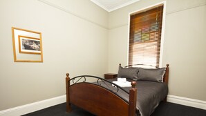 A bedroom with a wooden bed, a nightstand, a framed picture on the wall, and a window with blinds. - Peden's Hotel (Cessnock)