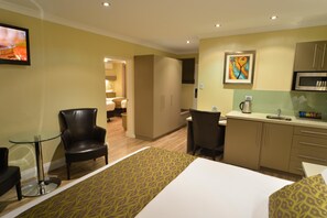 A hotel room with a bed, a desk, a chair, a kitchenette, and a bathroom visible through an open door.