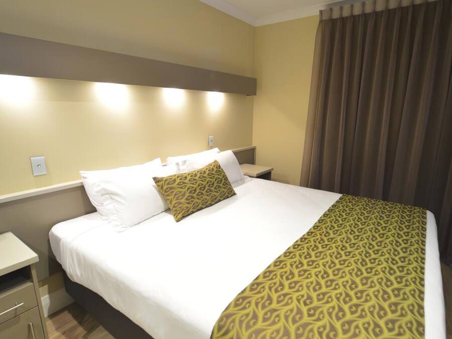 Una habitación de hotel con cama, mesita de noche, lámpara y ventana con cortinas.