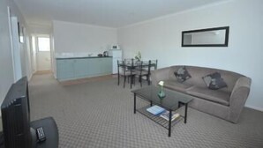 Sala de estar com sofá, mesa de centro e televisão. Há uma área de jantar com cadeiras e mesa. Uma cozinha é visível ao fundo.