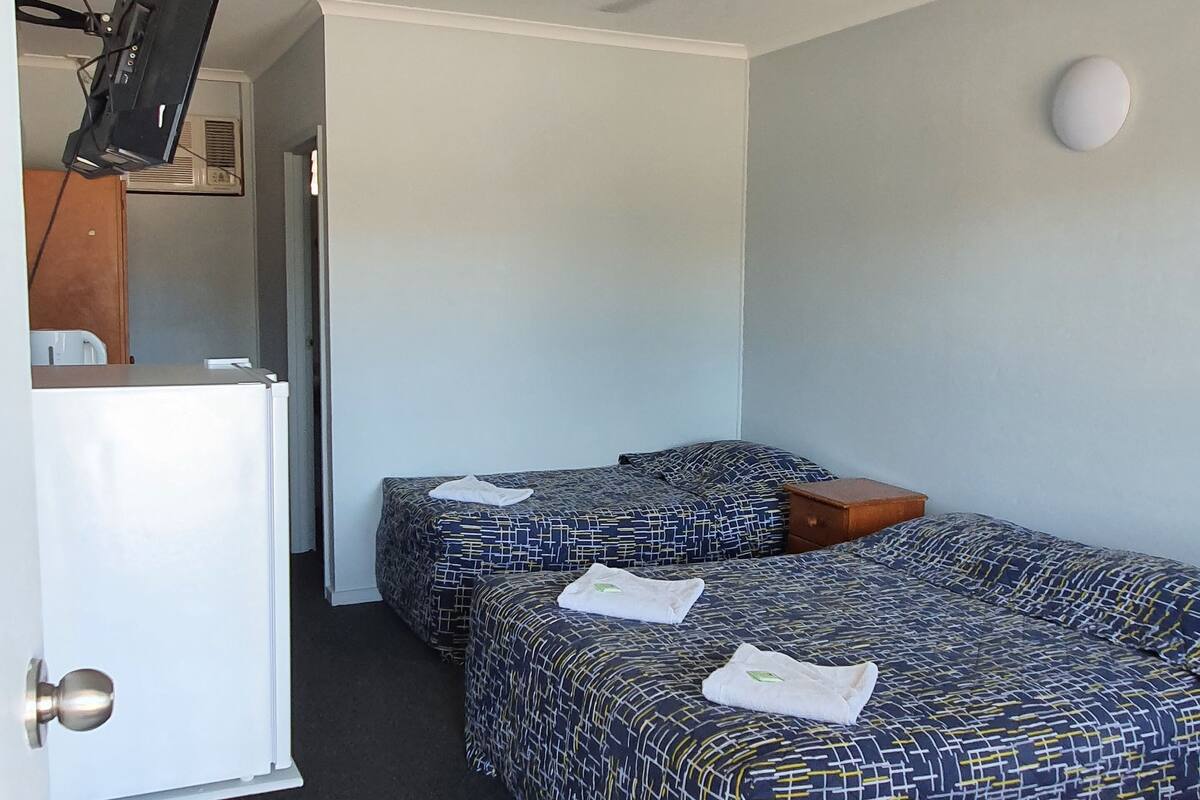 Una habitación con dos camas, un ventilador de techo, un televisor sobre un soporte y una puerta con picaporte blanco.