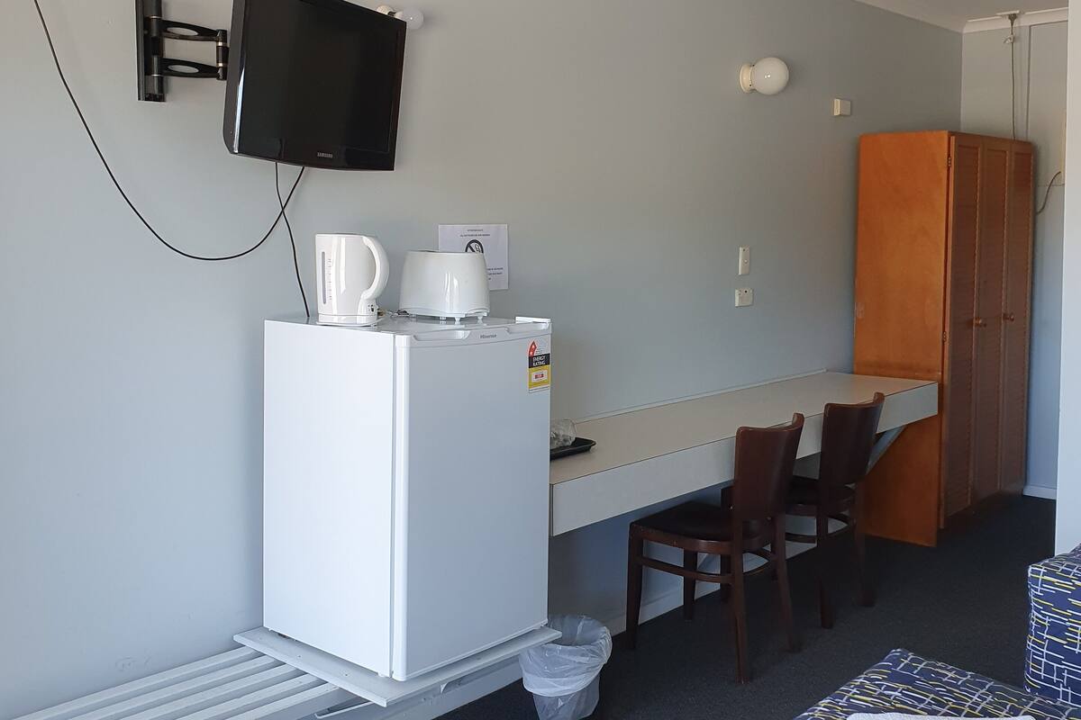 Habitación de hotel compacta con kitchenette, una cama individual, un escritorio con silla y ventilador de techo.