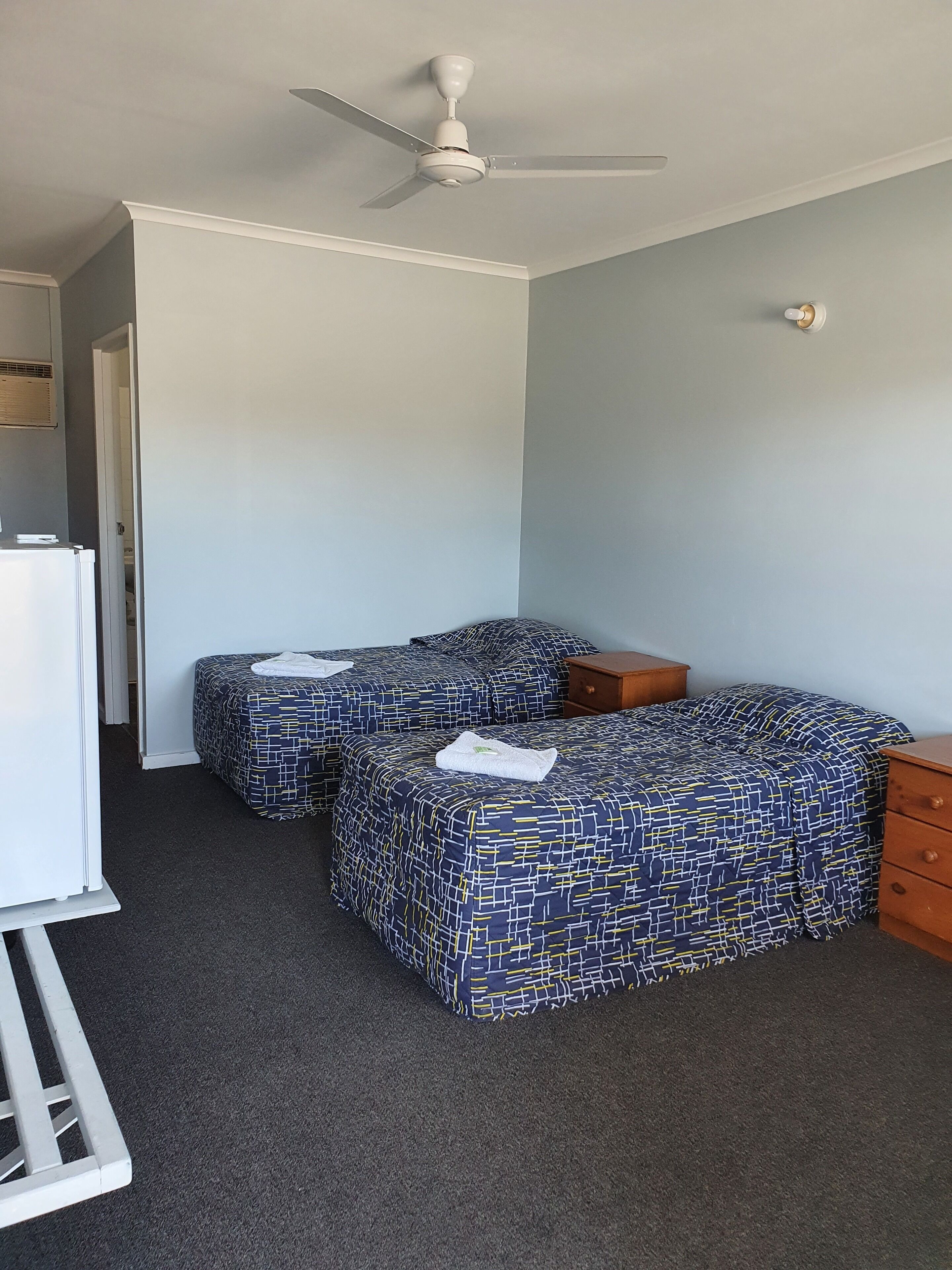 Une chambre avec deux lits, un ventilateur de plafond, un radiateur blanc et une table de chevet en bois.